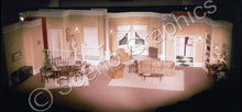 Load image into Gallery viewer, A stage set depicting a living room scene from a play, with a yellow sofa, a white fireplace, and a brown dining table. An example of The Odd Couple Set built by a high school using ScenoGraphics&#39; blueprints
